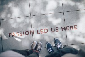 A photo of the pavement taken from above. Two people’s feet are visible in the shot. In front of their feet is text reading ‘passion led us here’.