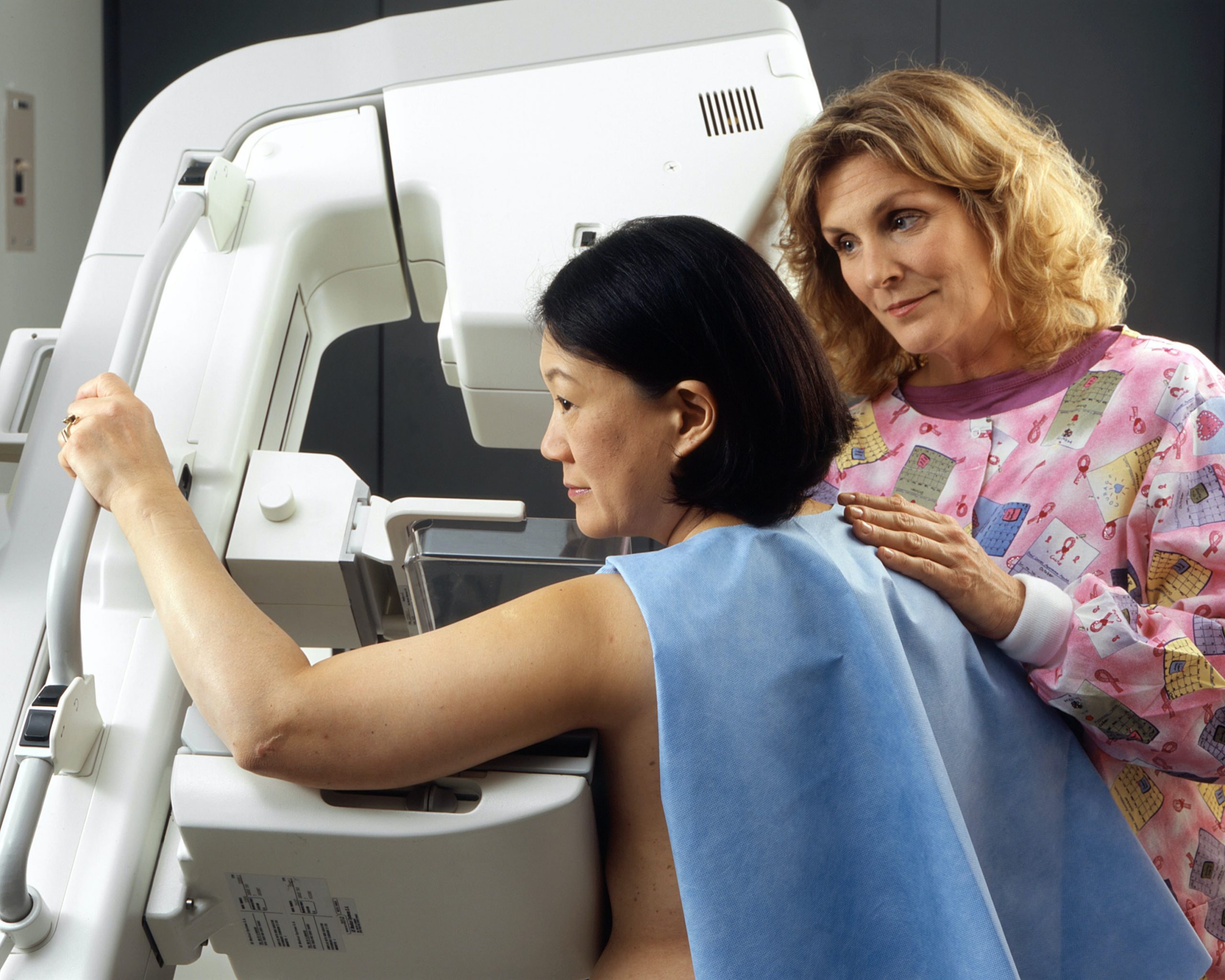 An Asian woman is having a mammogram, supported by a white female medical professional