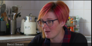 Becci is a white woman with short red and orange dyed hair. She is sat in her kitchen speaking to a reporter who is off camera.